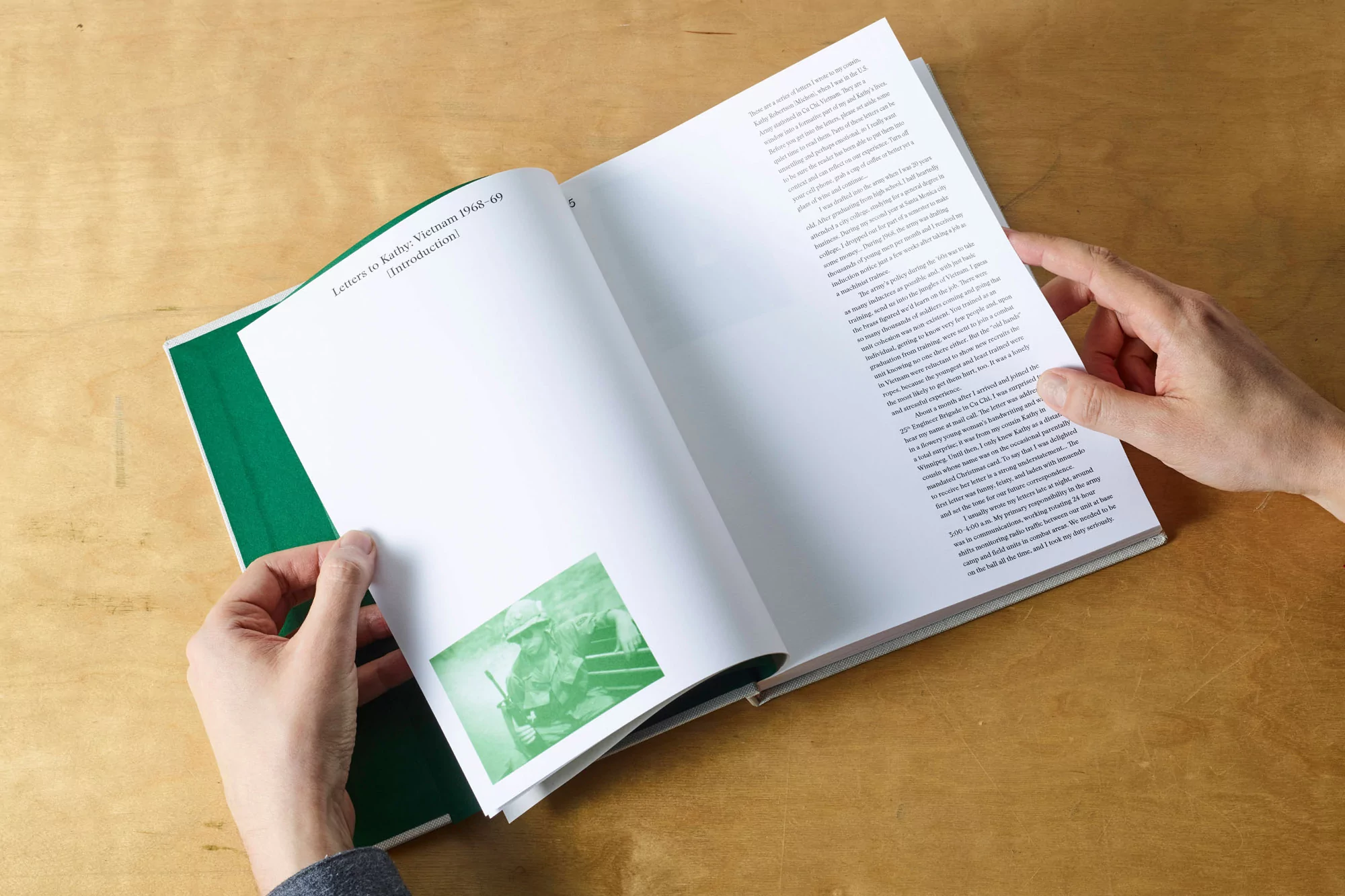 Hands holding an open book with green typography and a photographic insert on a wooden tabletop.