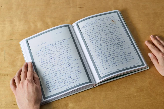 Open journal showing handwritten letters, accented by human hands. The neutral background emphasizes the blend of analog writing and personal touch.