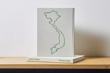 Grey hardcover book with a green outline of Vietnam standing upright on a wooden shelf next to a second book