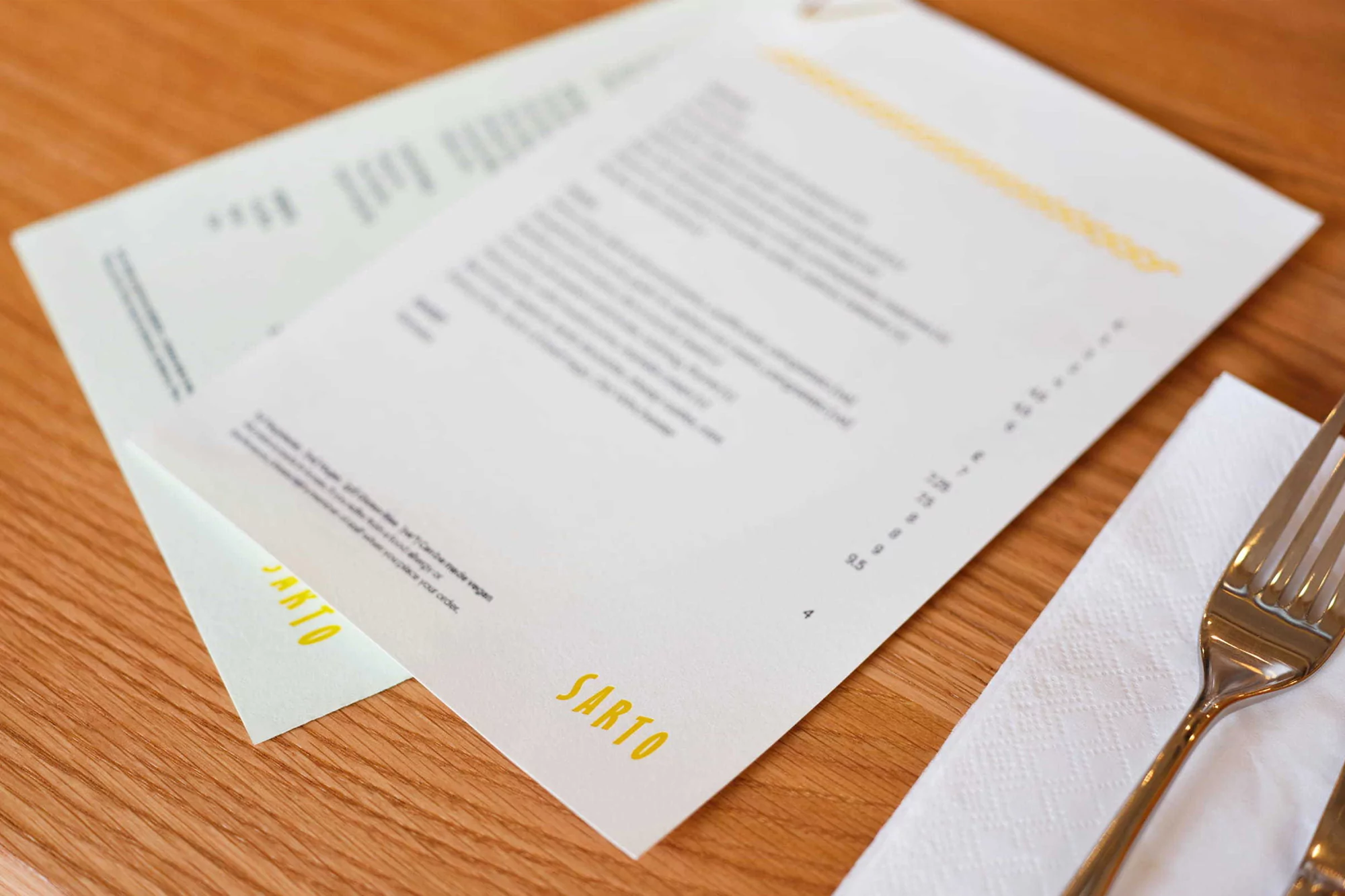 Restaurant menu on a wooden table next to cutlery and a white napkin, displaying dishes and prices at an indoor dining setting.