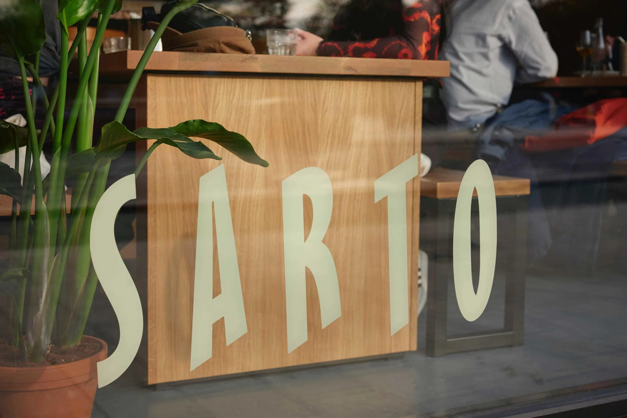 Potted plant near the window of a restaurant with the large white text 'SARTO' on the window; people sitting at tables inside.