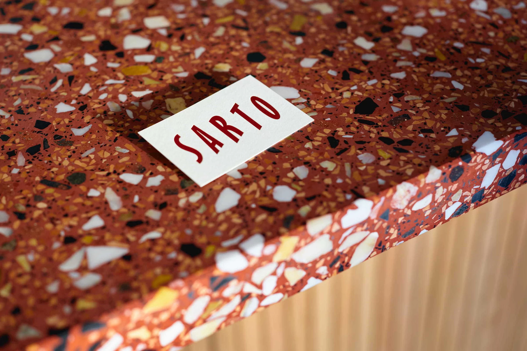 Close-up of a terrazzo countertop with red and multicolored stone chips, showcasing a white business card with the word 'SARTO' in red letters.