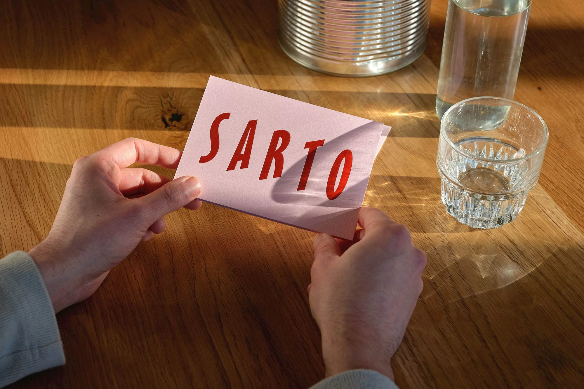 Close-up on hands holding pink card with red 'SARTO' text, clear drinkware, wood table, and metallic container in background.