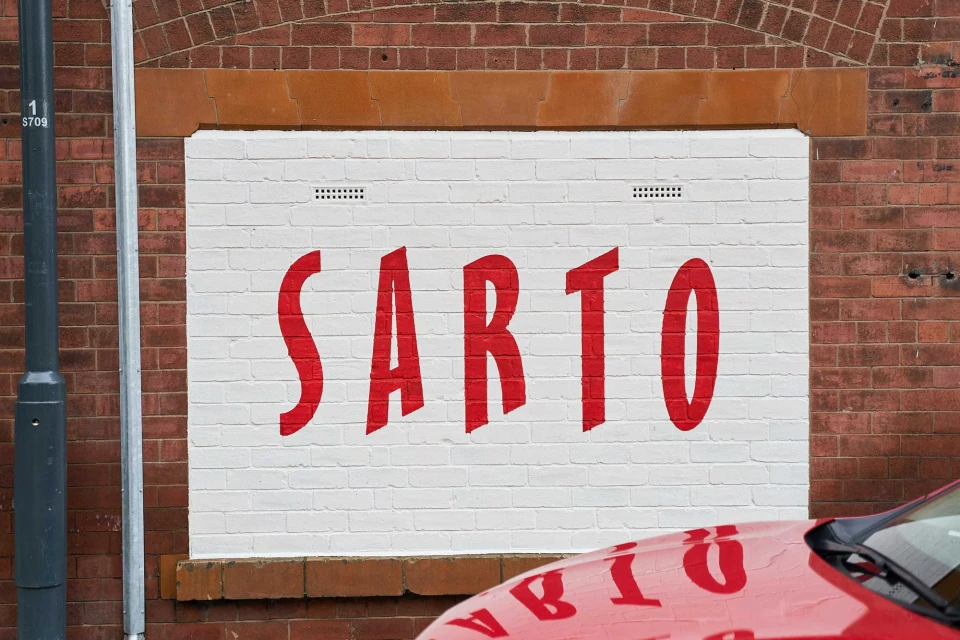 Red 'SARTO' sign painted on a white brick wall, with a glimpse of a red car in the foreground.
