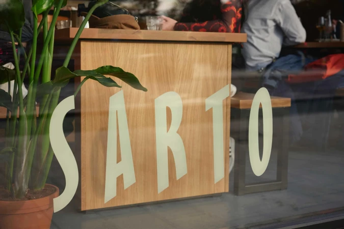 Window reflection showing large cream typography spelling SARTO on a wooden counter inside a restaurant