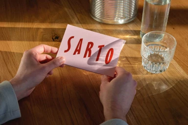Close-up of hands holding a pink card with red SARTO typography on a wooden table with glasses of water.