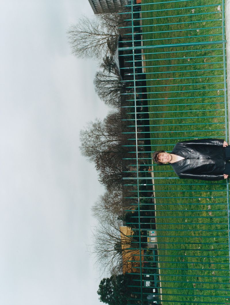 A person stands behind a green metal fence near a grassy field, with bare trees and a cloudy sky in the background on a gloomy day.