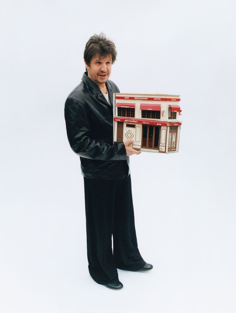 Man holds a detailed model of a European-style building in front of a seamless white background, dressed in a black jacket and pants.