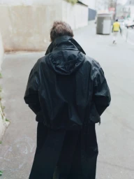 A person seen from behind walking down an urban street wearing a black hooded jacket and long black coat.