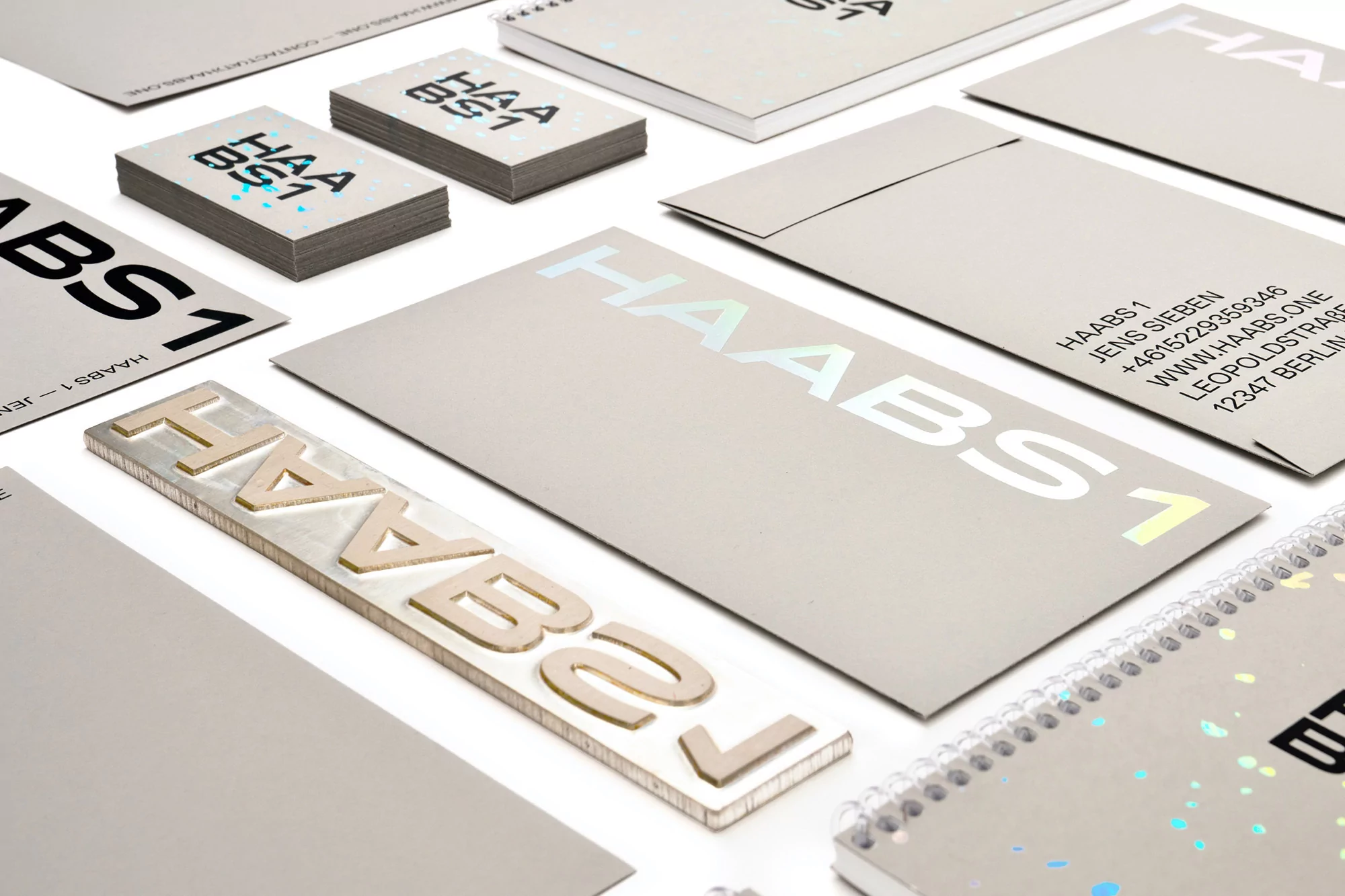 Arrangement of stationery with holographic details: business cards, envelopes, notebooks, and a printing block spelling 'HAABS'.