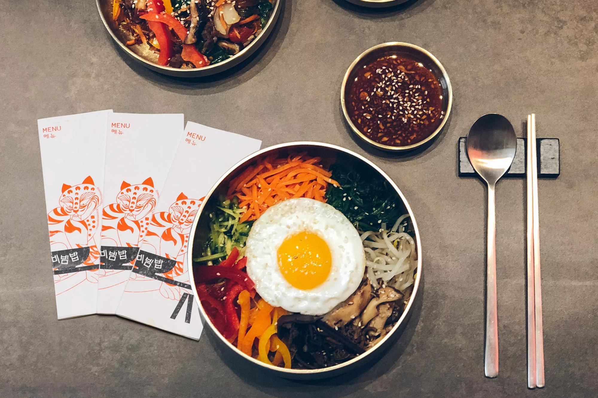 Overhead shot of Korean bibimbap in a brass bowl with an egg, chopsticks, spoon, sauce, and menus on a gray surface.