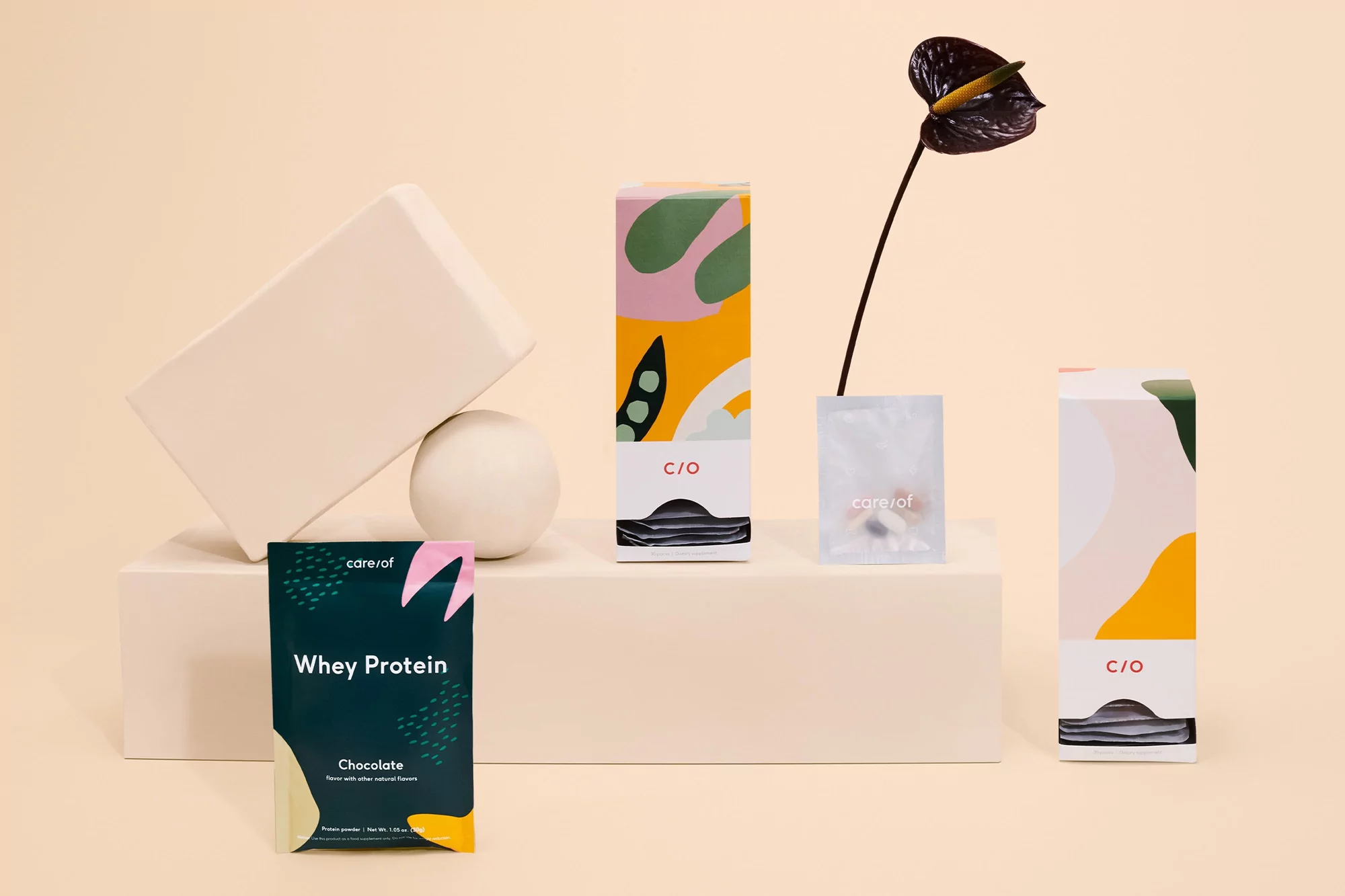 Still life shot of Care/of supplement packets in colorful packaging with geometric shapes and a deep purple Anthurium flower.