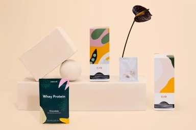 Product packaging display for REMAINS featuring colorful boxes, whey protein packets, and a dark flower against a cream background.