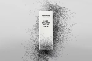 White product box for REMAINS post-trimmer beard balm centred over scattered black hair clippings on light grey seamless background