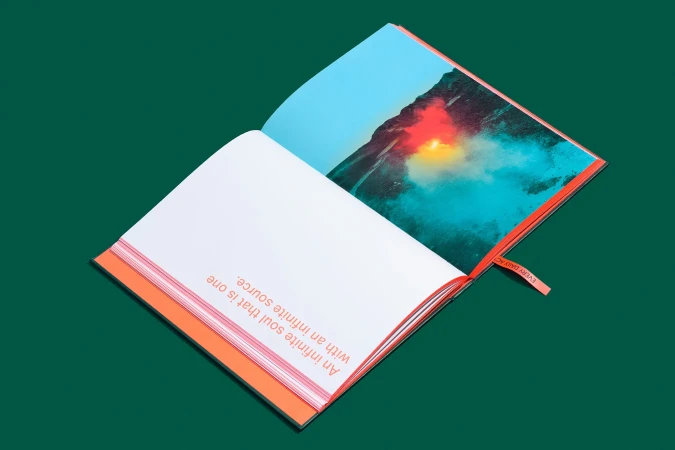 Open book on green surface. Left page has text, right page displays colorful mountain landscape with red and blue tones. Orange details.