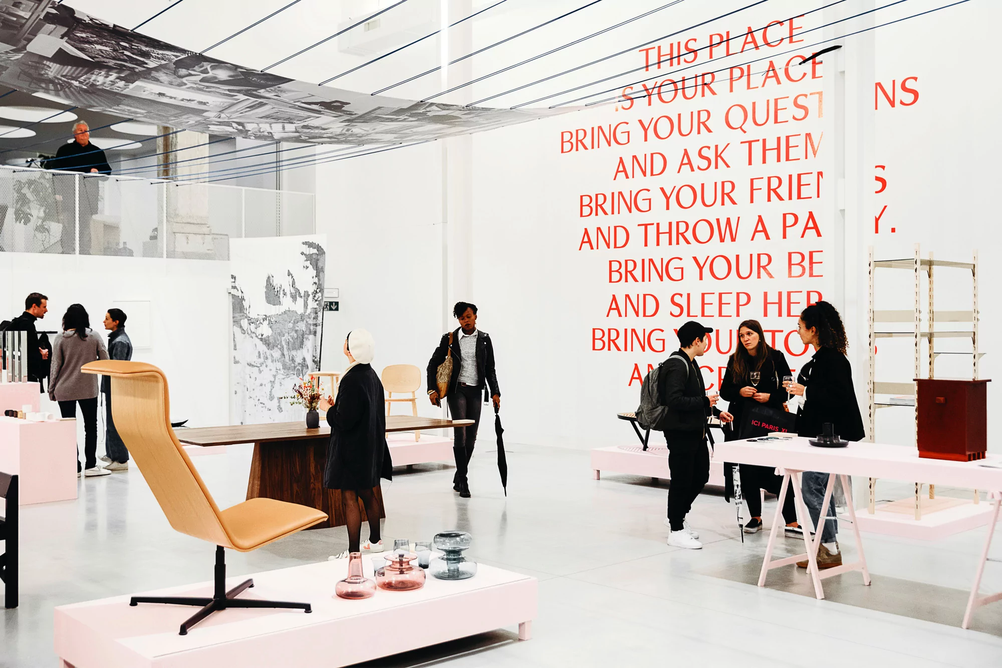 Art installation with people inside a gallery featuring furniture and large red text on the wall. The space is well-lit and modern.