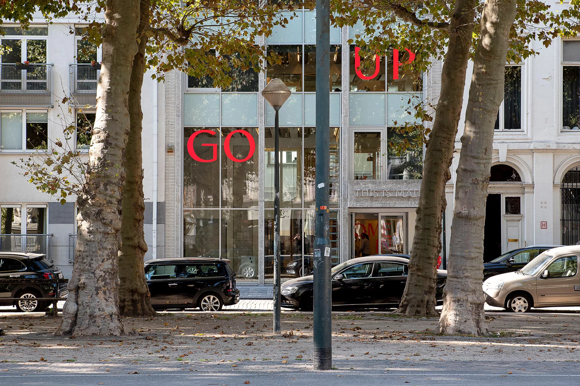 Exterior view of a modern building with large windows displaying red 'GO UP' lettering, framed by trees and cars on the street.
