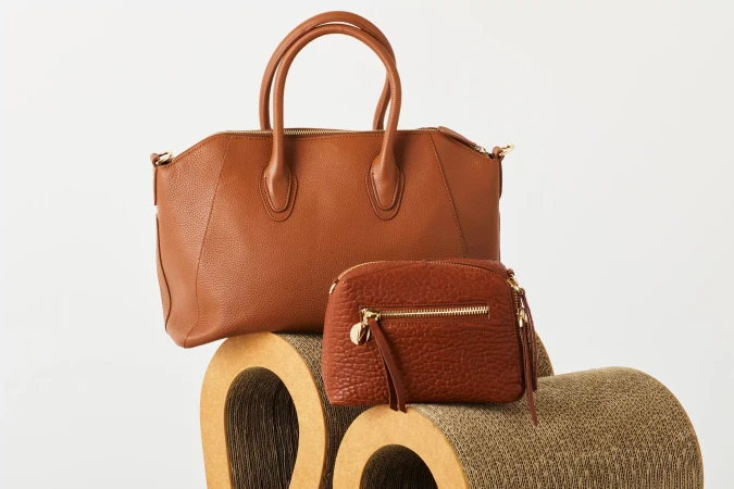 Two brown leather handbags, a satchel and a smaller purse, are displayed on a textured, tan paper sculpture against a plain white background.