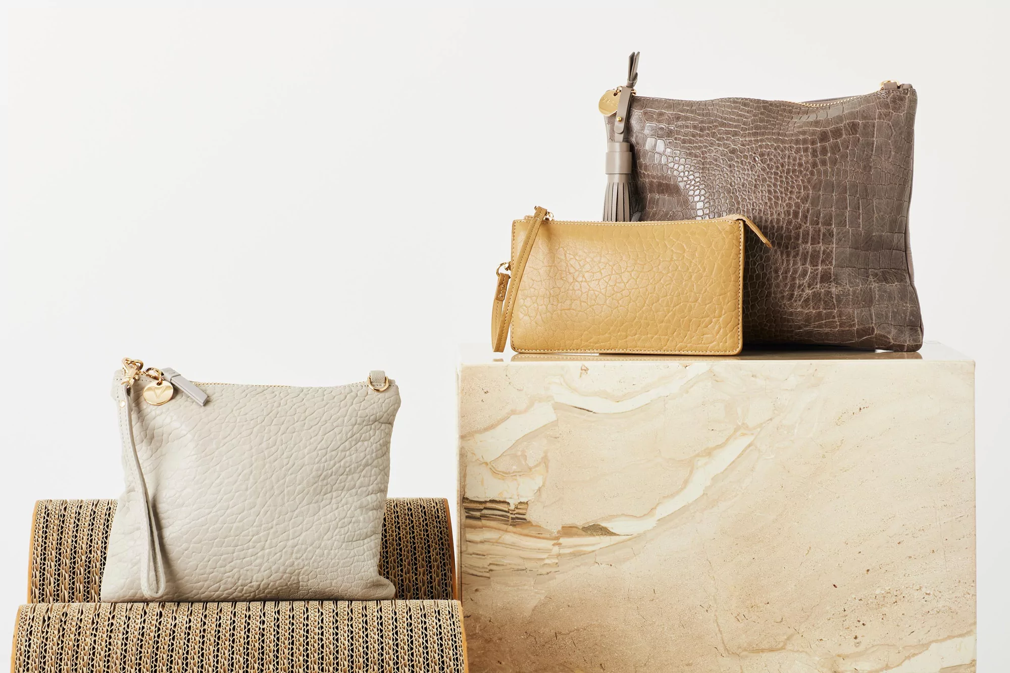 Three textured leather handbags in neutral tones are staged on a marble plinth and corrugated surface against a bright white background.