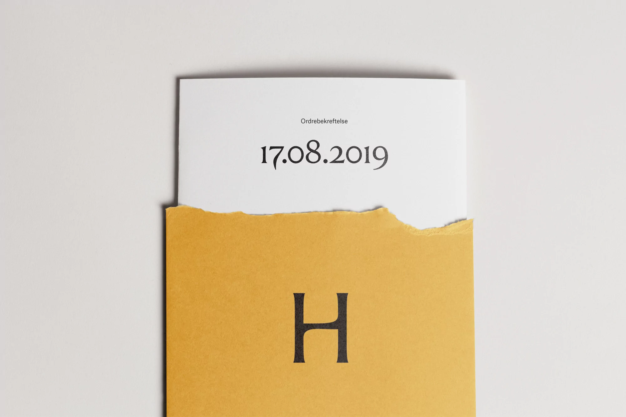 Close-up of a stationery design featuring a white sheet with the date 17.08.2019 partially covering an ochre paper with letter H.