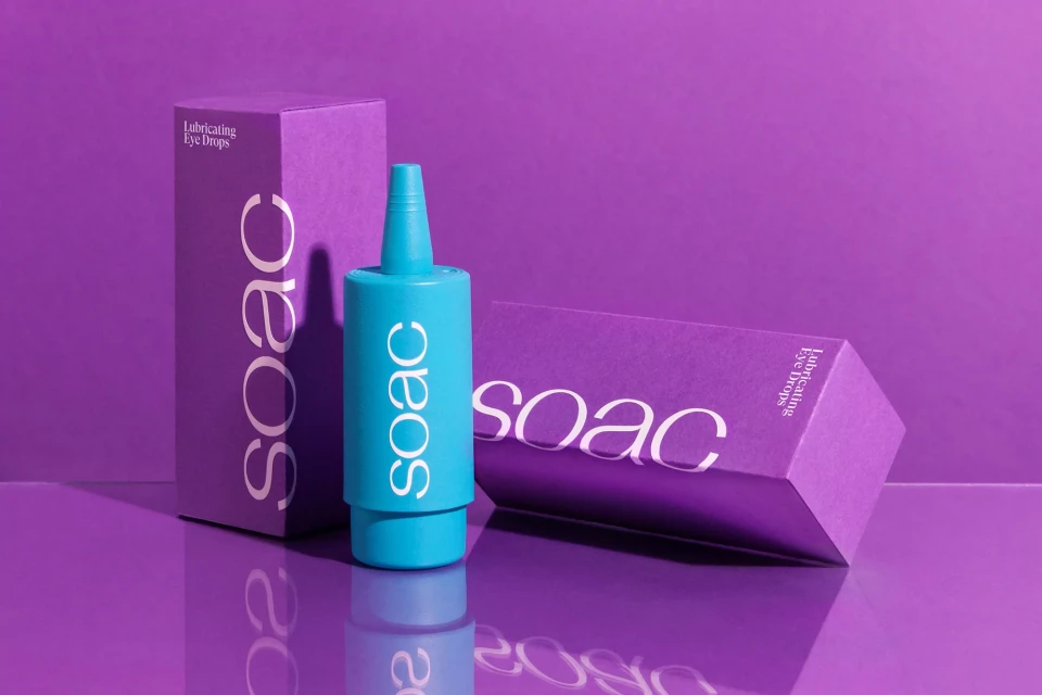 A turquoise bottle of eye drops sits next to purple product packaging, reflecting on the shiny purple surface against a solid purple backdrop.
