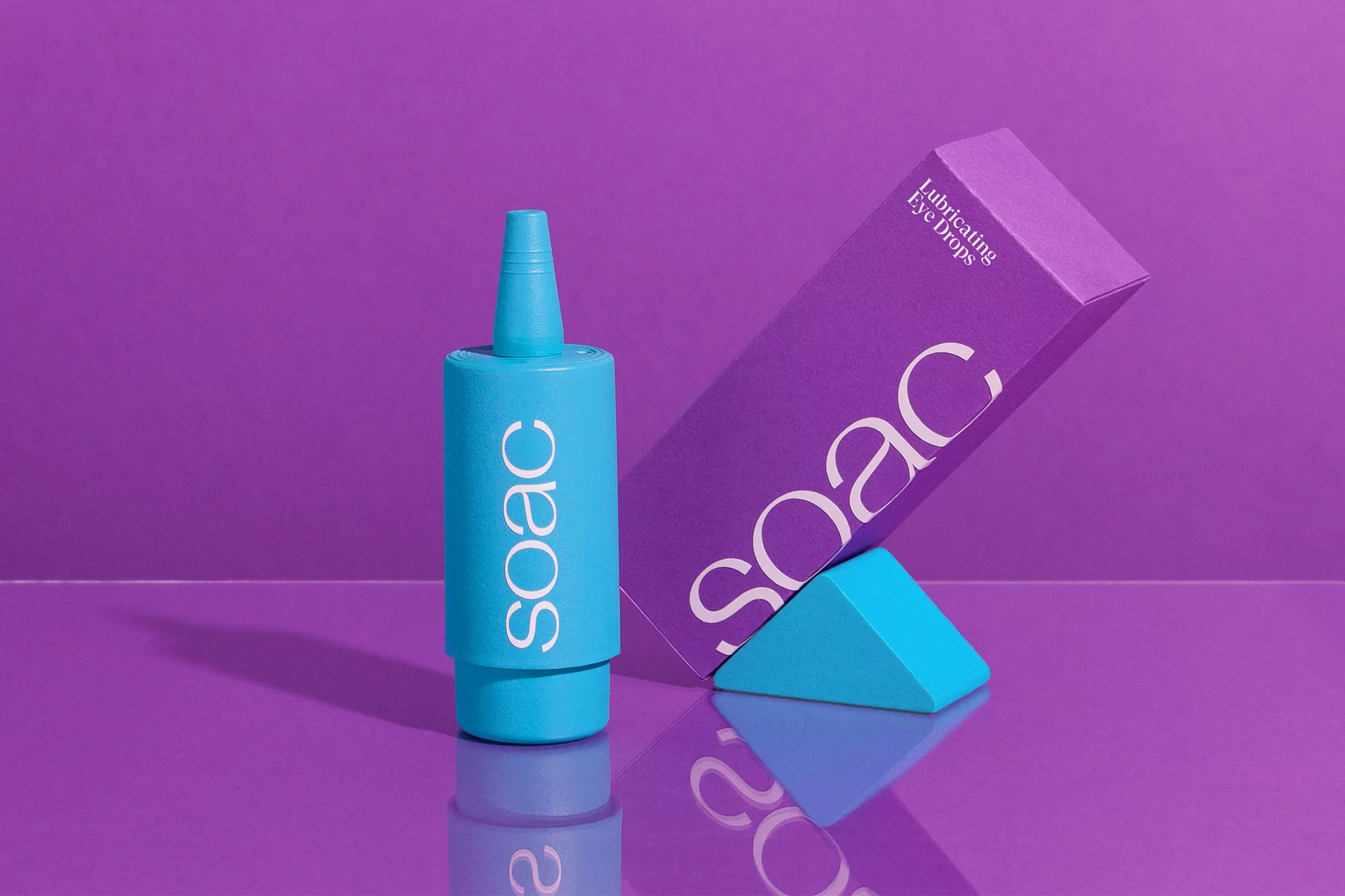 Turquoise eye drop bottle with white text beside a purple box of eye drops leaning on a turquoise triangle on a purple surface.