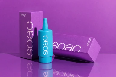 Bright blue eye drops bottle standing between two purple packaging boxes on a reflective surface