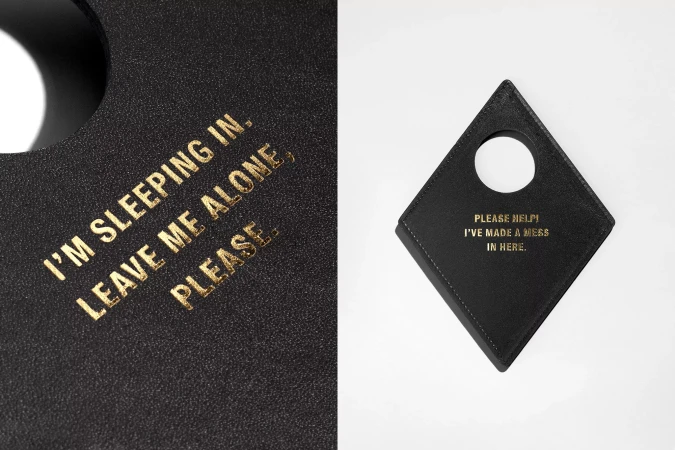 Close-up of two black leather door hangers with gold text. One reads 'I'm sleeping in, leave me alone, please', the other 'Please help! I've made a mess in here'.