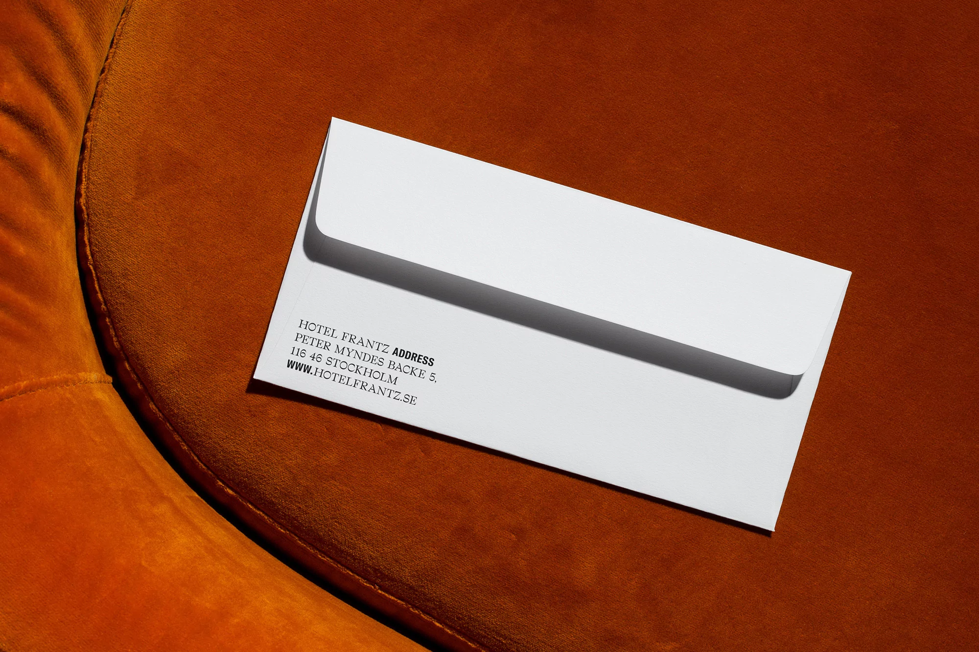 White envelope featuring Hotel Frantz address lies on burnt orange textured fabric in an eye-level studio shot, emphasizing textures.