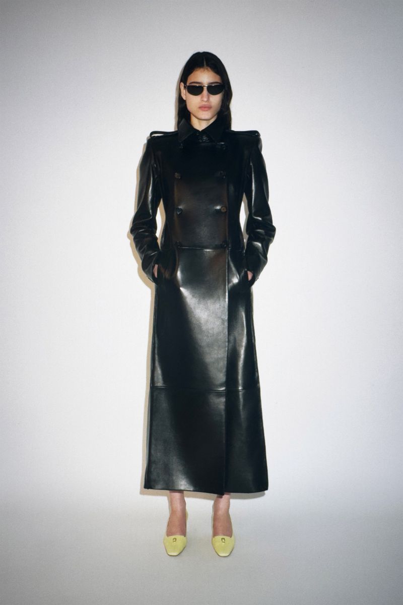Fashion photo of a woman wearing a black leather coat and sunglasses against a white background, showcasing a high-fashion look.