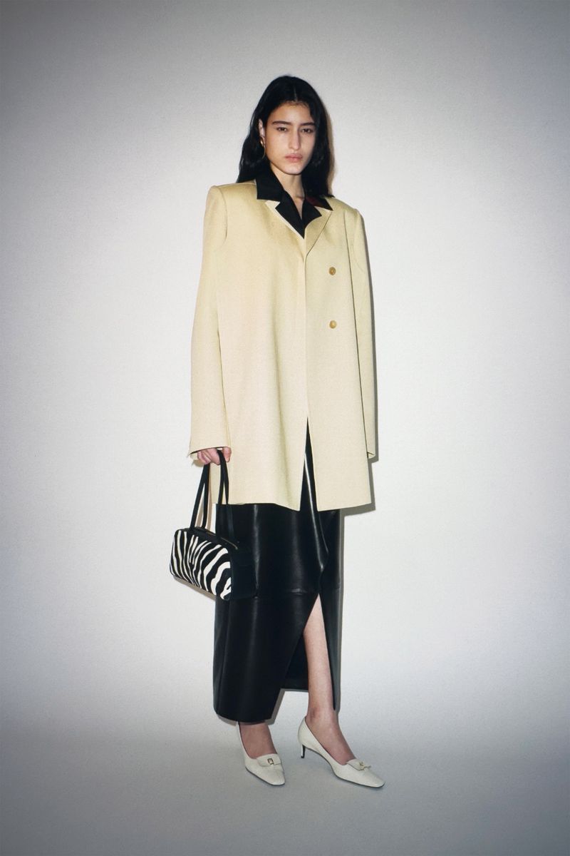Fashion model poses in studio wearing cream overcoat, black dress, zebra bag, and kitten heels against a neutral backdrop in a studio shot.