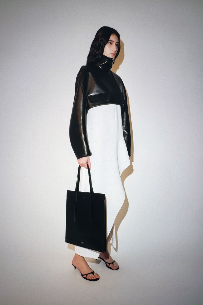 Fashionable woman in black jacket and white dress poses indoors with a black tote bag, showcasing modern style against a light background.