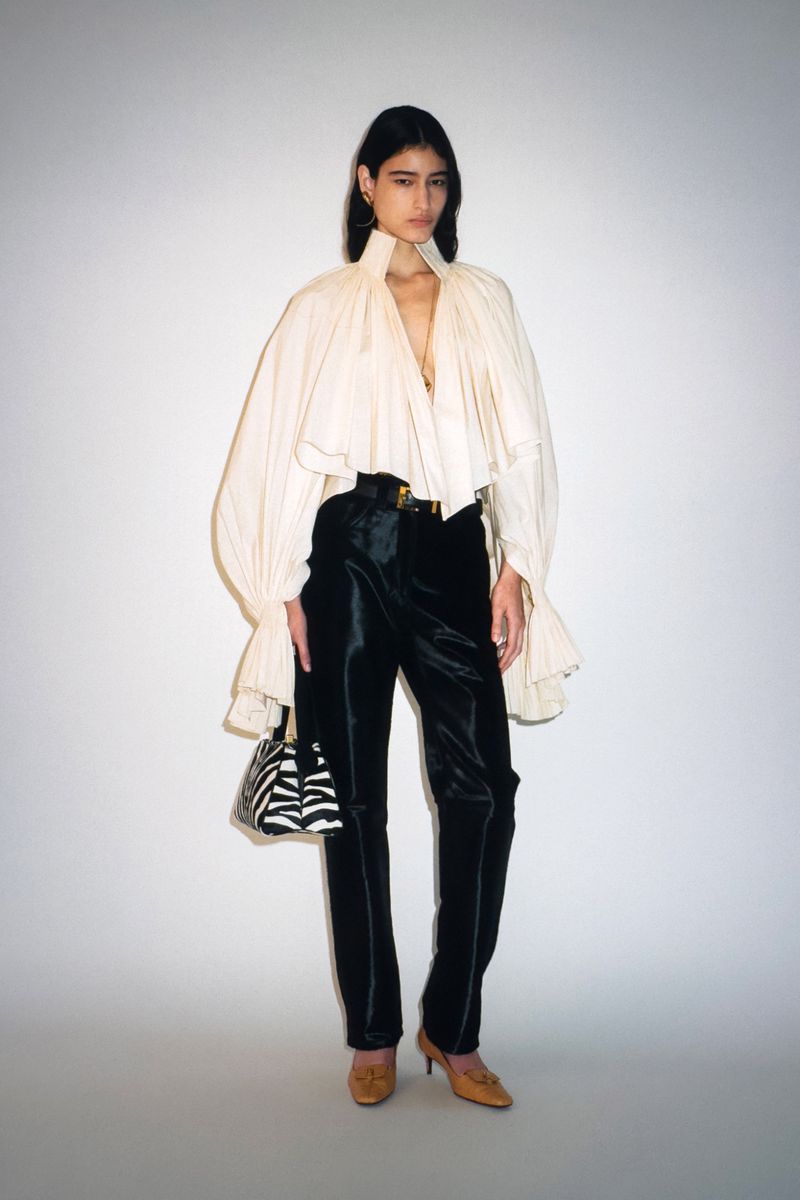 Fashion model poses in a white, oversized blouse with black velvet pants and a zebra-print handbag in a studio setting with neutral backdrop.