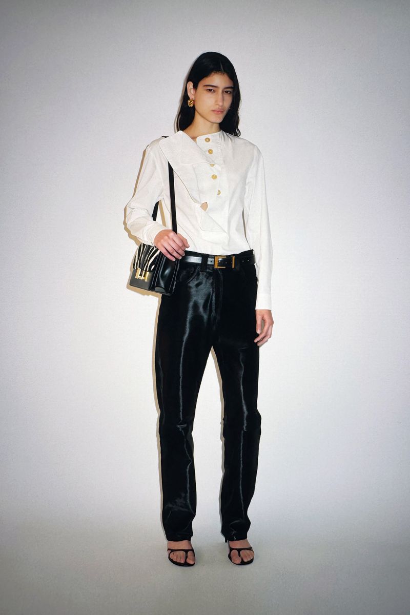 Elegant woman in a white blouse and black pants poses in a studio with a shoulder bag, capturing a high-fashion look.