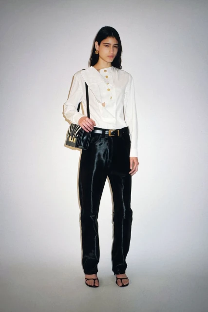 Elegant woman in a white blouse and black pants poses in a studio with a shoulder bag, capturing a high-fashion look.