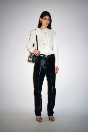 Elegant woman in a white blouse and black pants poses in a studio with a shoulder bag, capturing a high-fashion look.