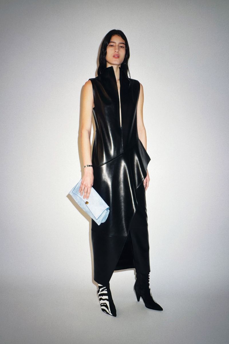 A stylish woman with long dark hair wearing a black sleeveless dress and zebra-print boots, holding a light blue clutch bag indoors.