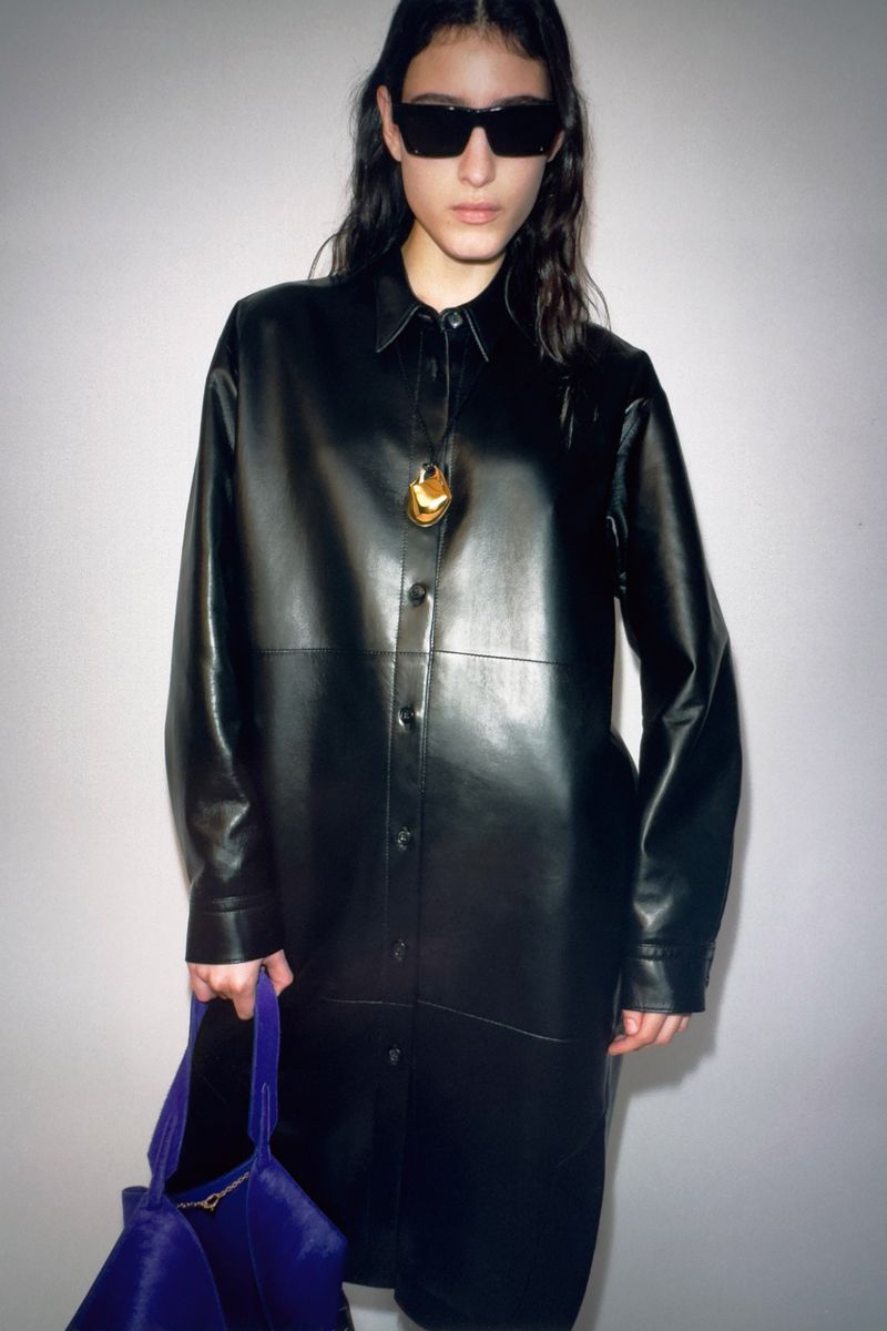 Fashionable young adult woman in long-sleeved leather dress, sunglasses, gold pendant necklace, and carrying purple tote bag.