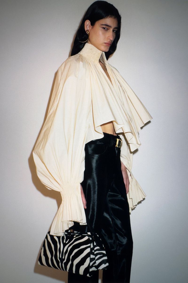 Adult female fashion model poses in voluminous cream-colored blouse with black pants and zebra-striped handbag against white wall.