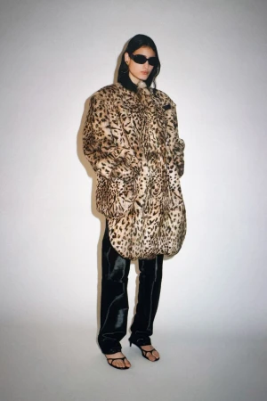 Woman in oversized leopard print coat, black pants, and sunglasses standing against a white background in a fashion forward pose.