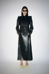 Model wearing a long black coat and dark sunglasses standing against a plain white studio background.