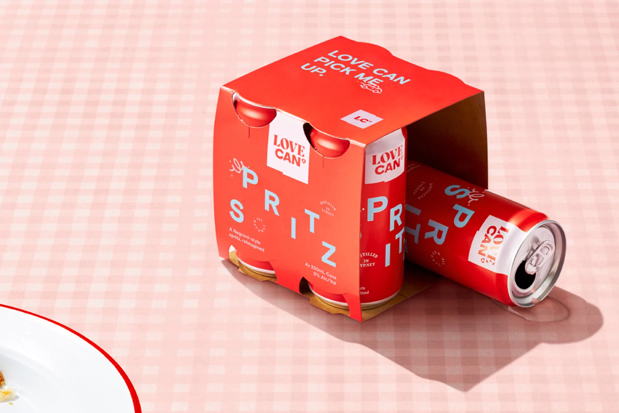 A pink gingham tablecloth with a 4-pack of Love Can Spritz cans with one can laying outside the packaging casting a shadow.