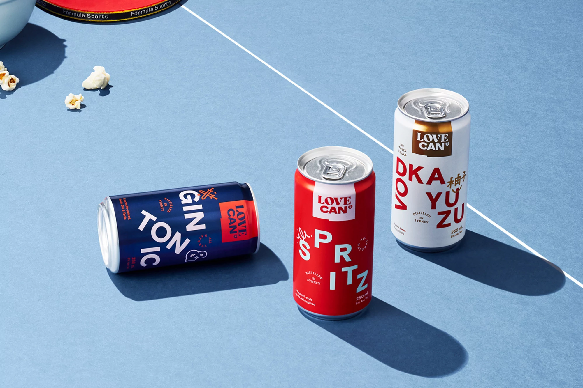 Three cans of Love Can brand sparkling alcoholic beverages: Gin & Tonic, Spritz, and Yuzu, displayed on a blue surface with scattered popcorn.