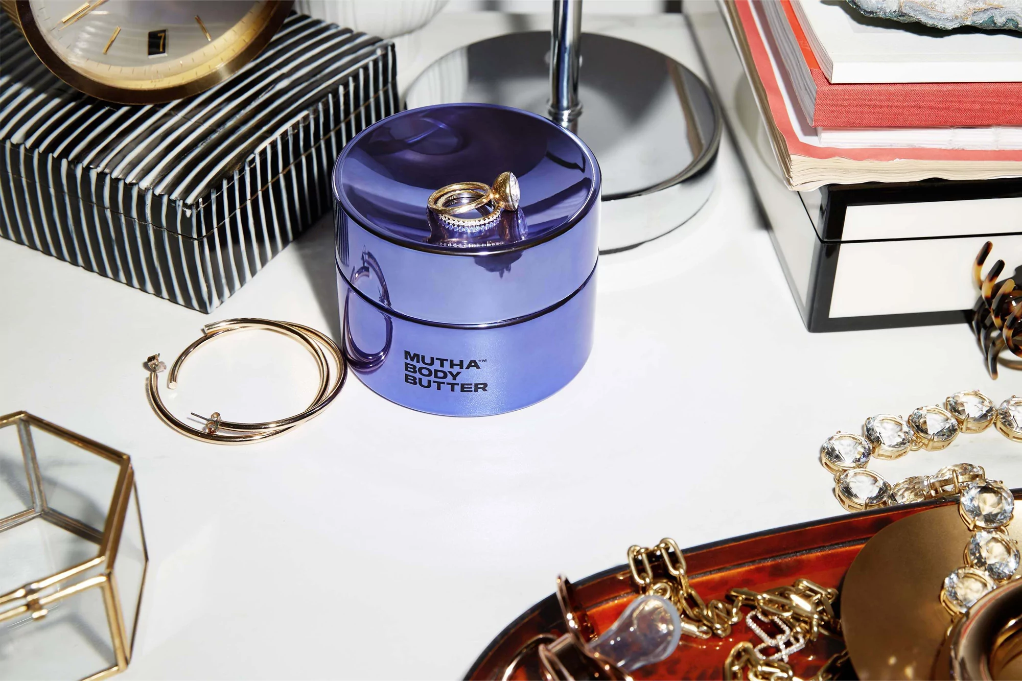 Close-up of a purple jar of Mutha Body Butter with jewelry on a white surface, alongside accessories and beauty products, indoors.