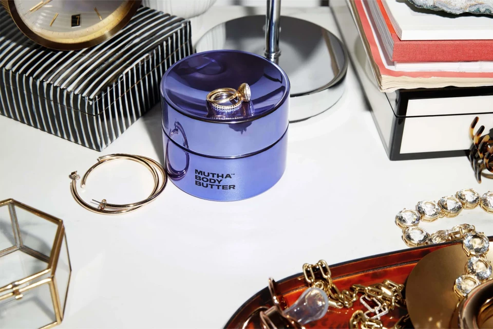 Close-up of a purple jar of Mutha Body Butter with jewelry on a white surface, alongside accessories and beauty products, indoors.