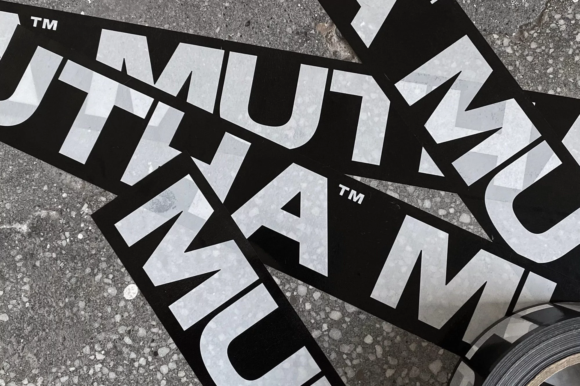 Overhead shot of black stickers with repeating 'MUTNAM' text in white on a grey concrete surface with a roll of black and white tape.