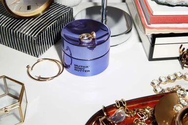 MUTHA body butter jar in glossy purple resting on a white tabletop surrounded by jewellery and books