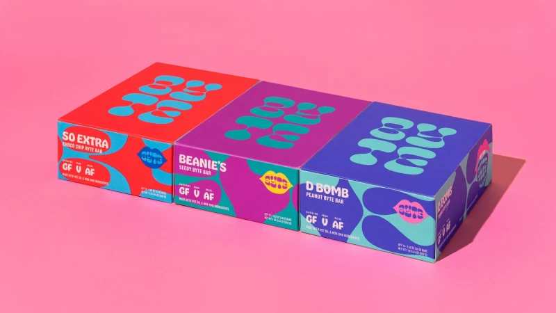 Eye-level studio shot of three colorful snack bar boxes: So Extra Choco Chip, Beanie's Seedy, and D Bomb Peanut Byte against pink backdrop.