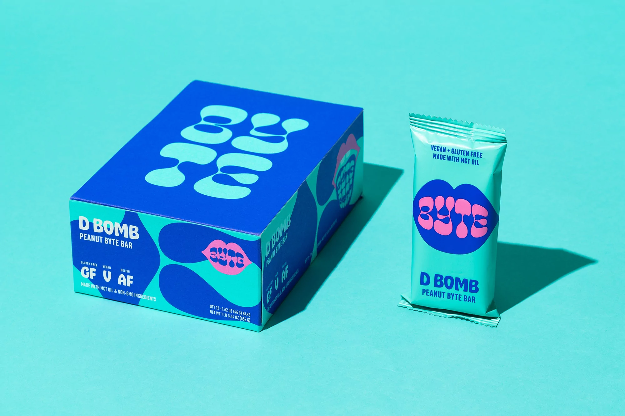 Box and individual package of D Bomb Peanut Byte Bar. Teal package has logo with a pink lip motif. Vegan, gluten-free snack.
