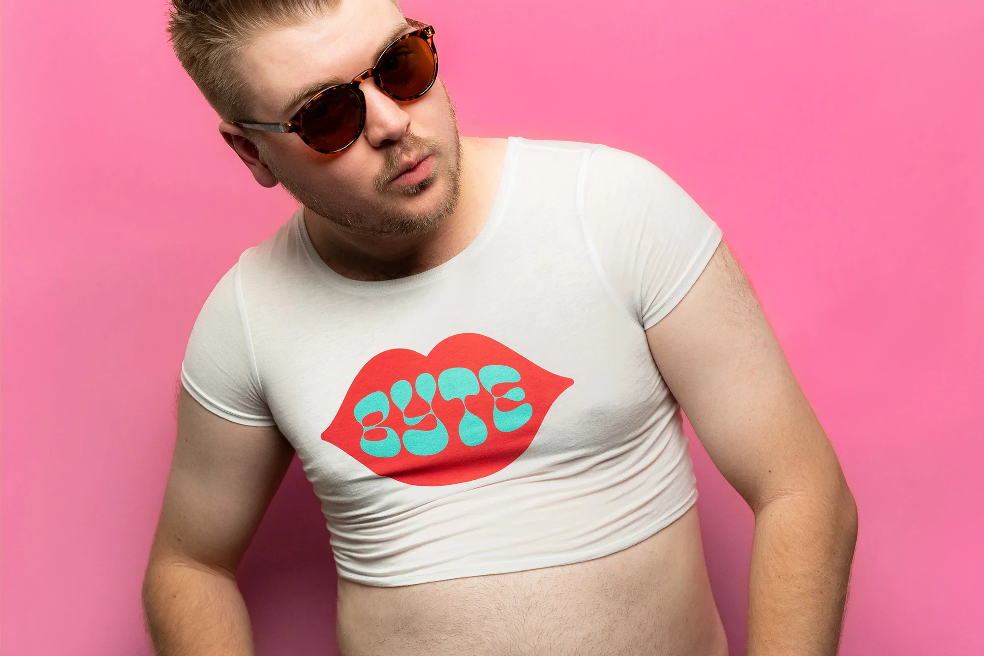 A blond adult male wears a white crop top with 'BITE' on it, sunglasses, looking left against a pink background in a medium shot.